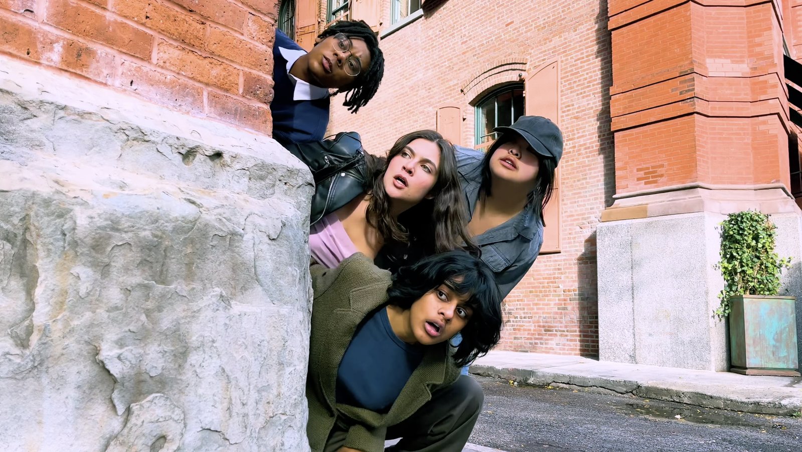 Four young adults peek warily around a stone corner on a New York street