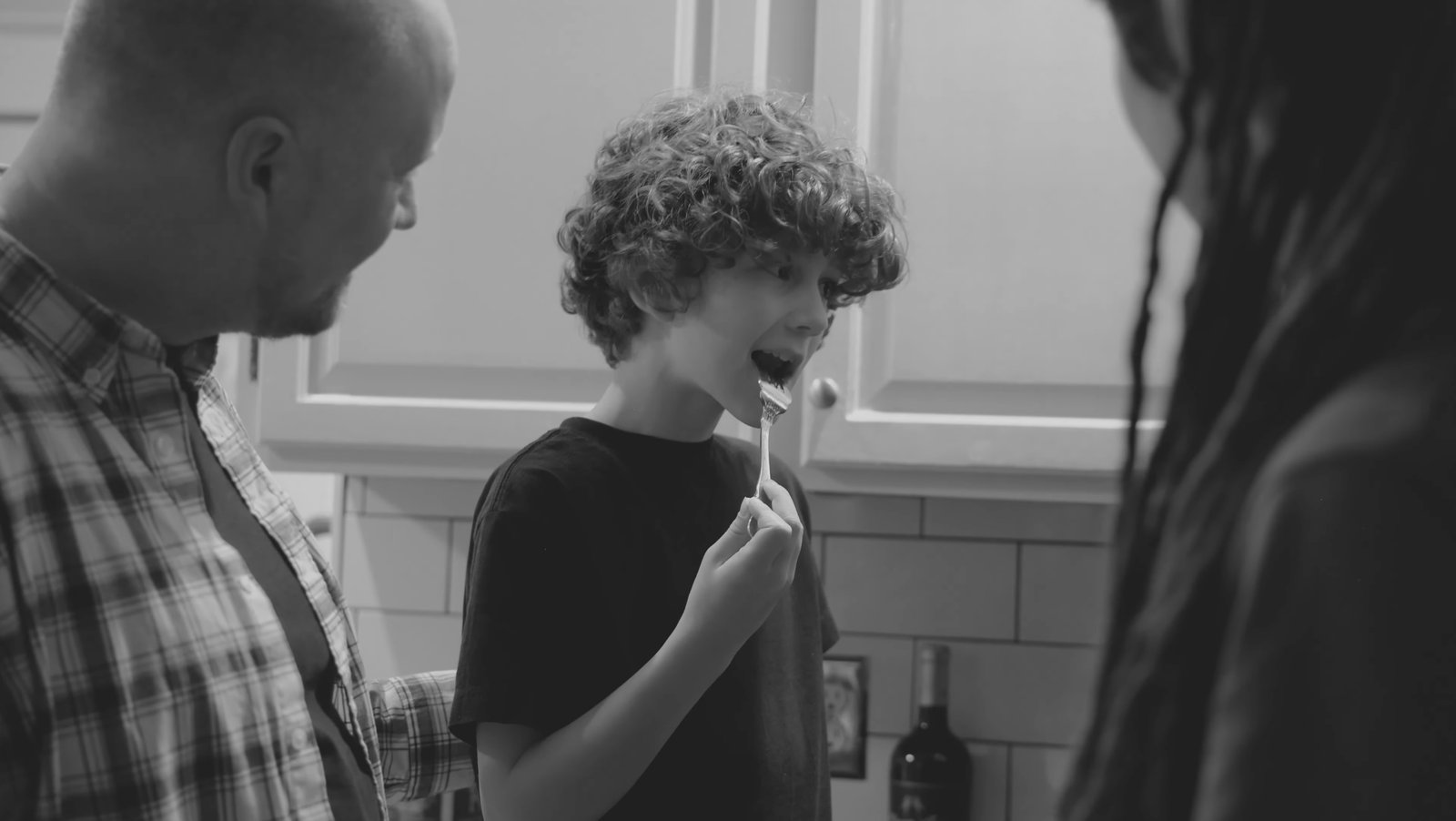 Black-and-white kitchen scene with a father, a son eating, and a mother