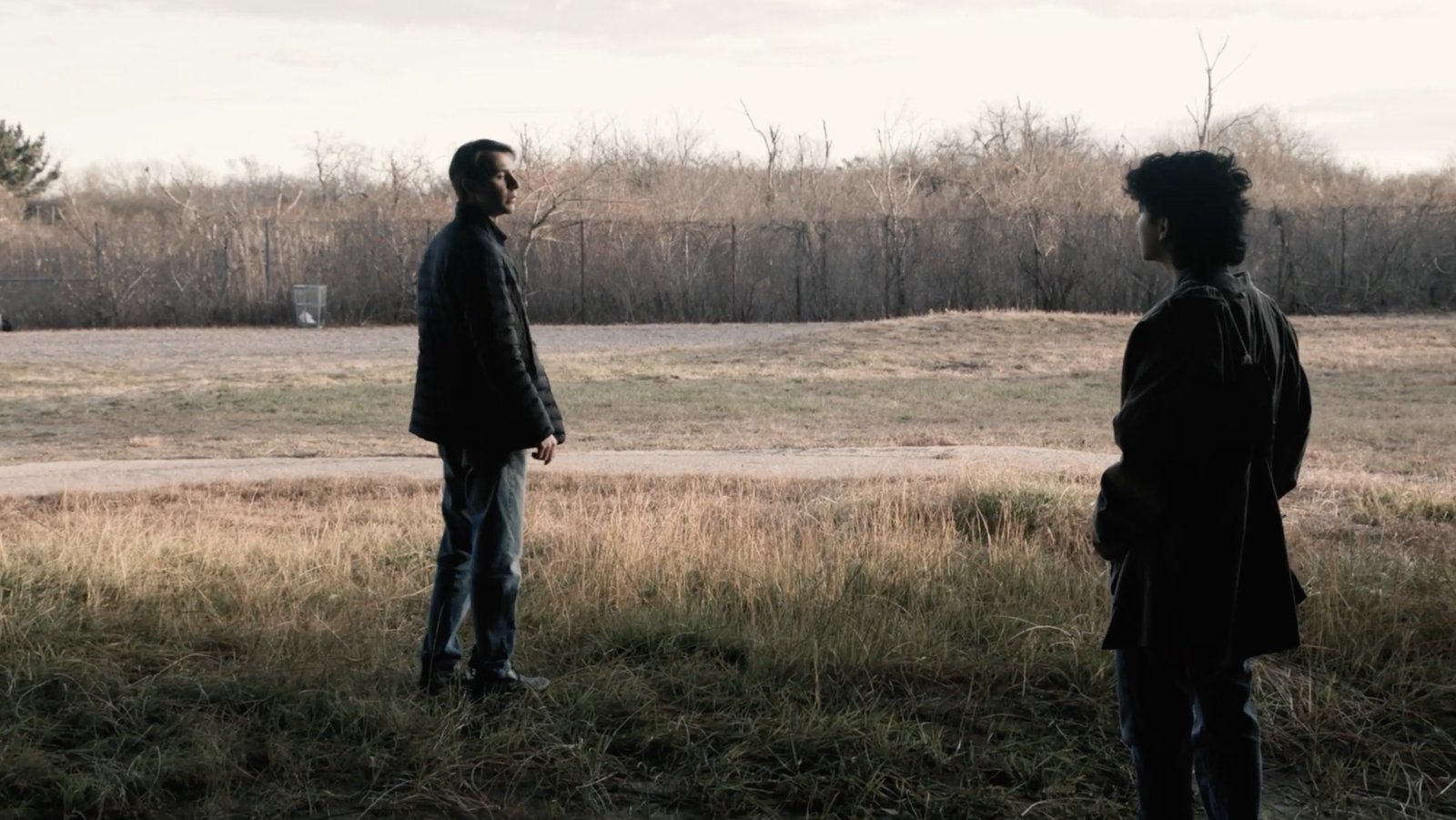 Two men stand facing each other across a desolate field of dry grass under an overcast sky